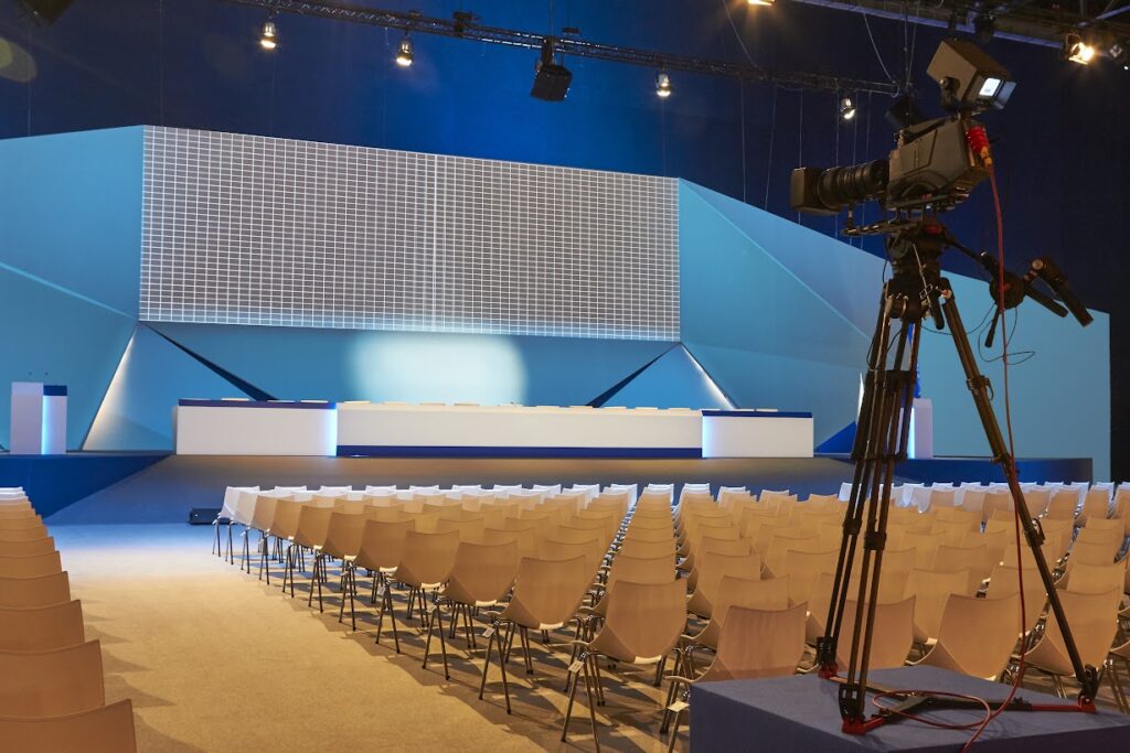 Una sala convegni allestita nell'ambito di una fiera: sullo sfondo, c'è il tavolo dei relatori. In primo piano, le sedute del pubblico viste da dietro e una telecamera.